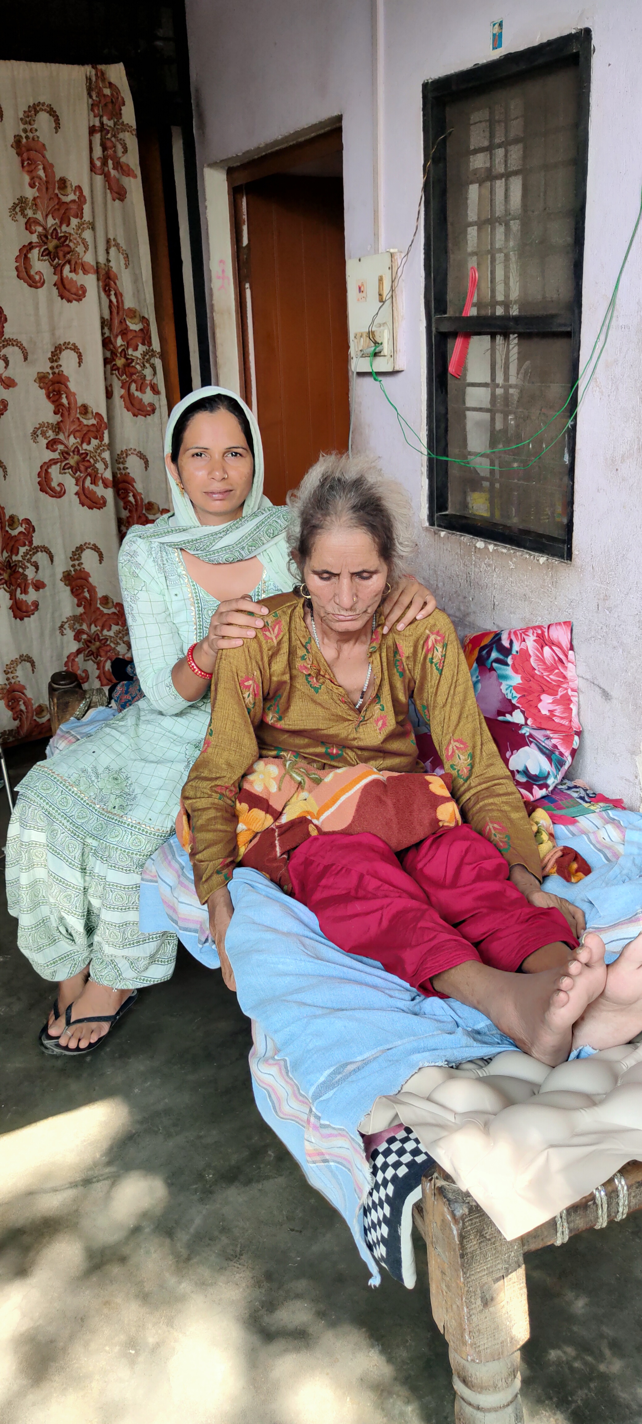 Dadi seated on veranda charpai with daughter's hands on shoulders